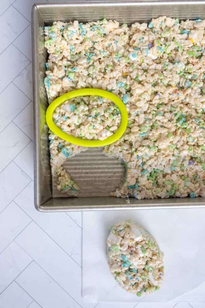 A pan of rice crispy treats with colorful sprinkles; an oval-shaped cutter is used to cut out a piece, which is placed on parchment paper nearby.