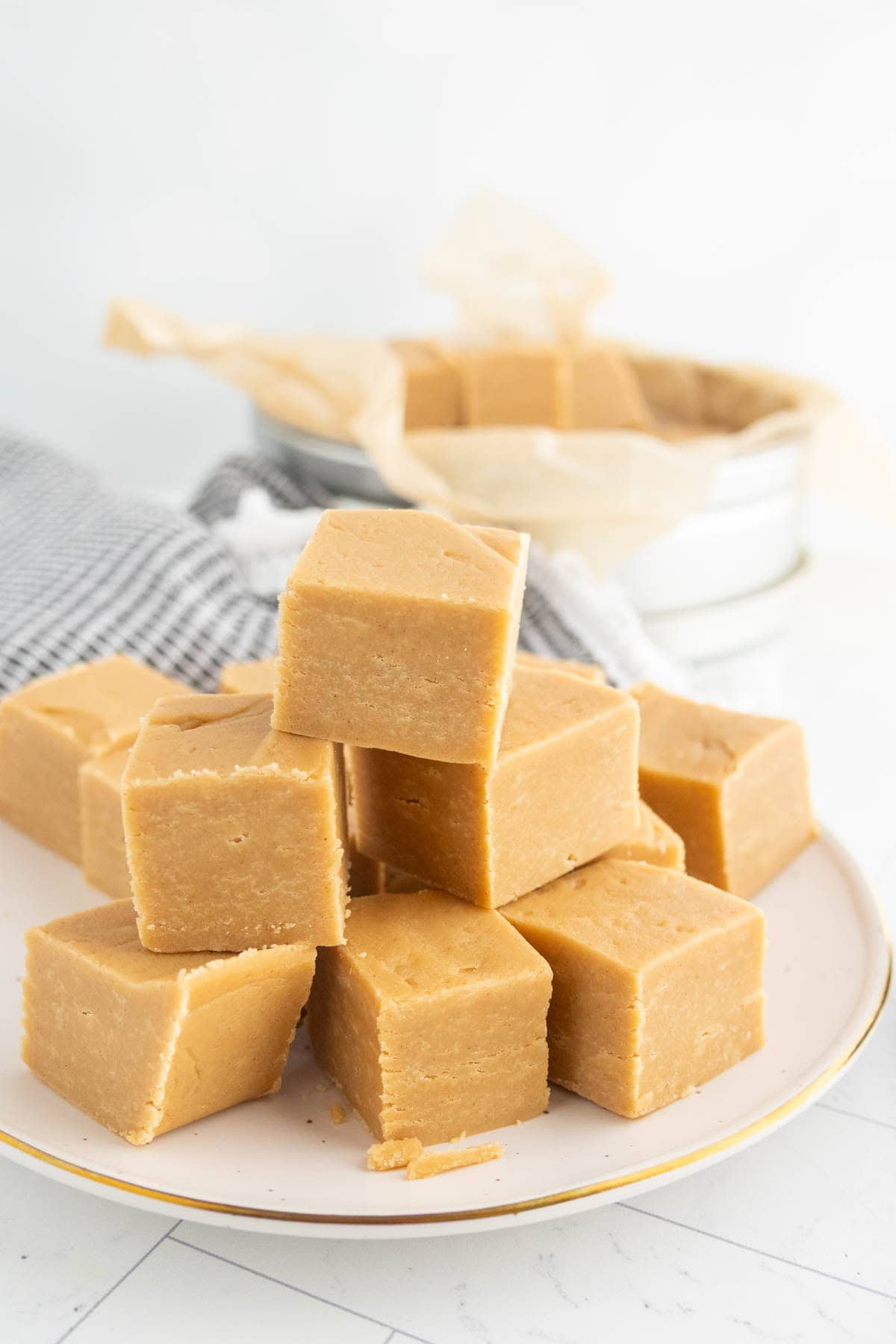 A plate with neatly stacked cubes of light brown fudge, with more fudge pieces and a cloth napkin in the background.