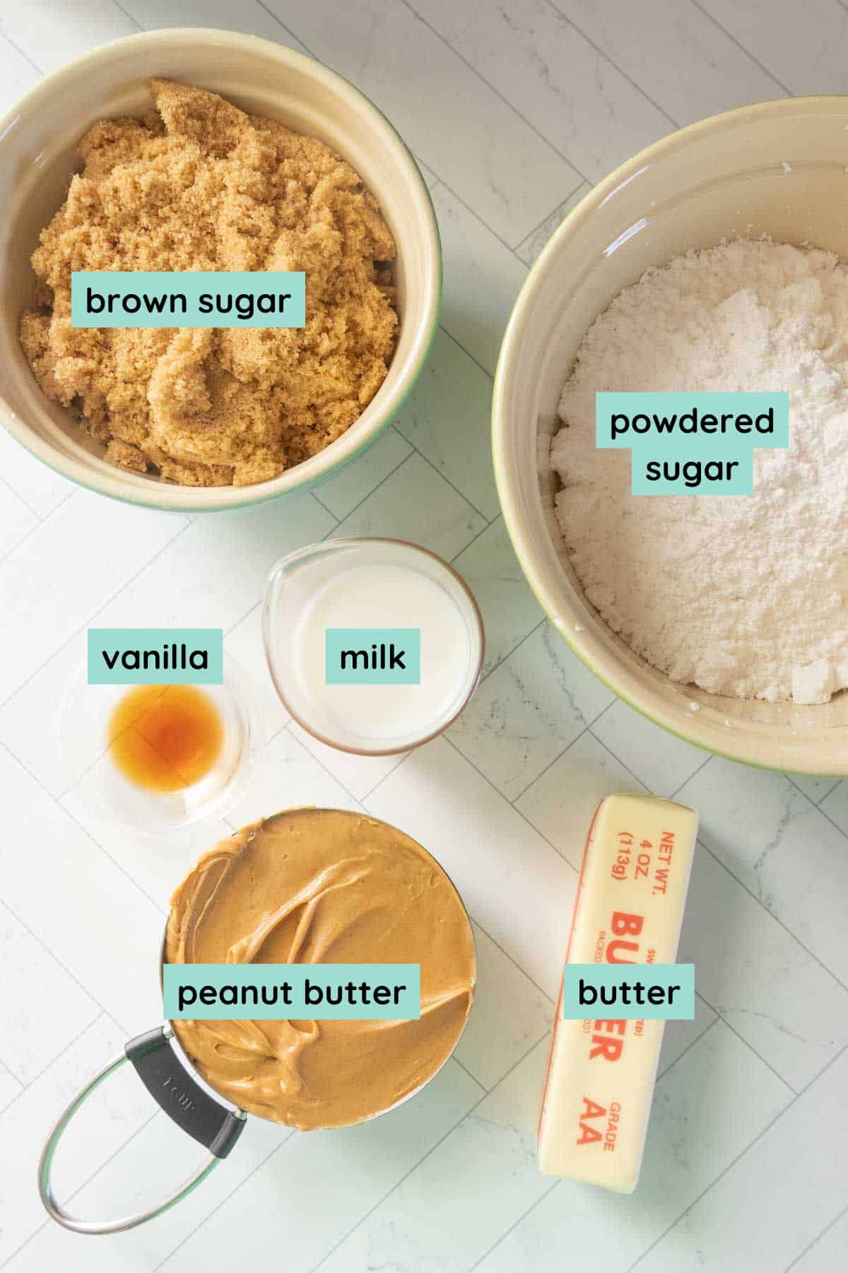 Bowls of brown sugar, powdered sugar, peanut butter, and butter, with small containers of vanilla and milk on a marble surface. Each ingredient is labeled.