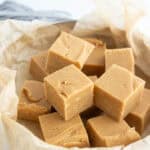 Squares of light brown fudge are stacked on parchment paper in a round metal tin.