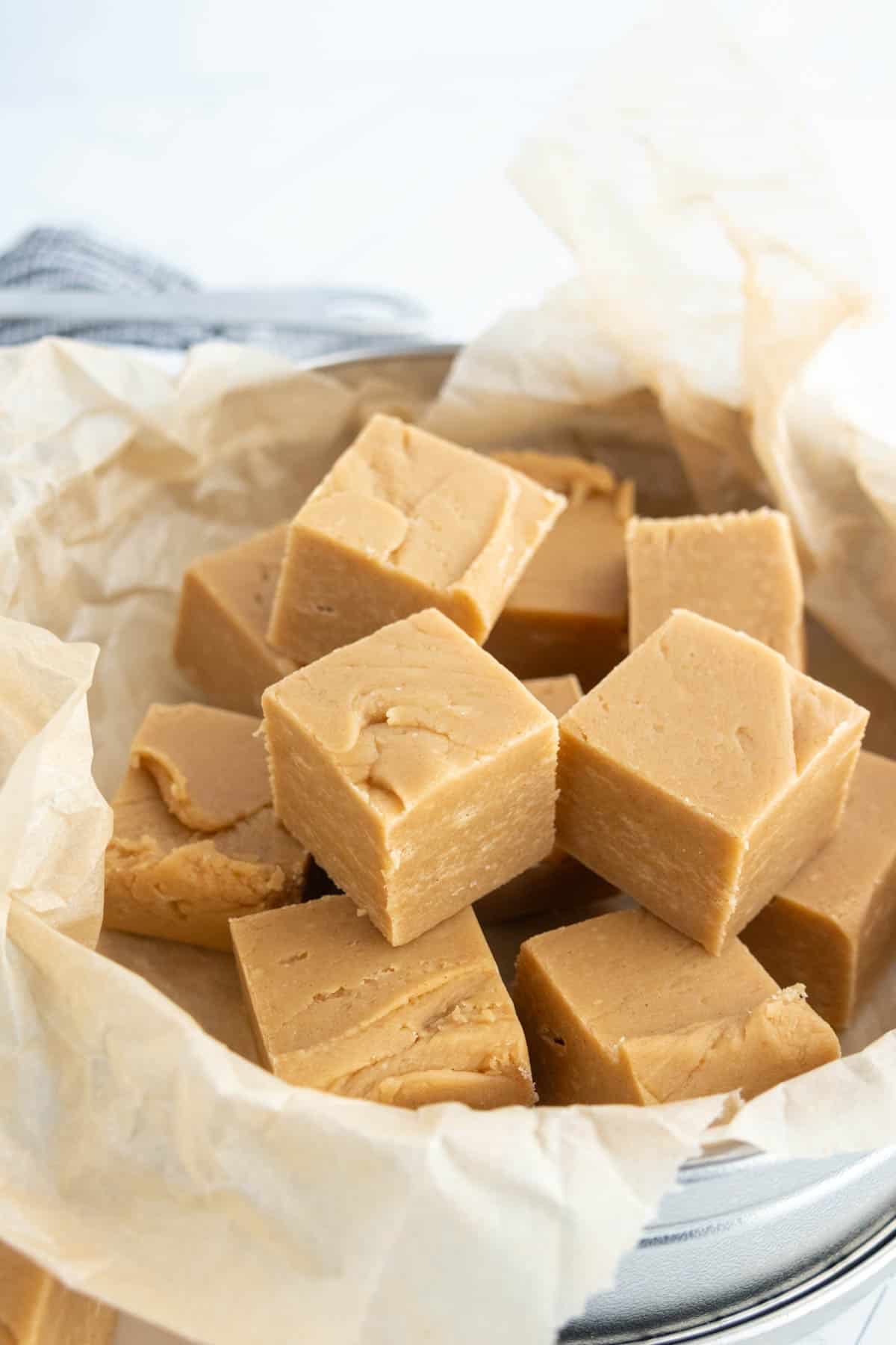 Squares of light brown fudge are stacked on parchment paper in a round metal tin.