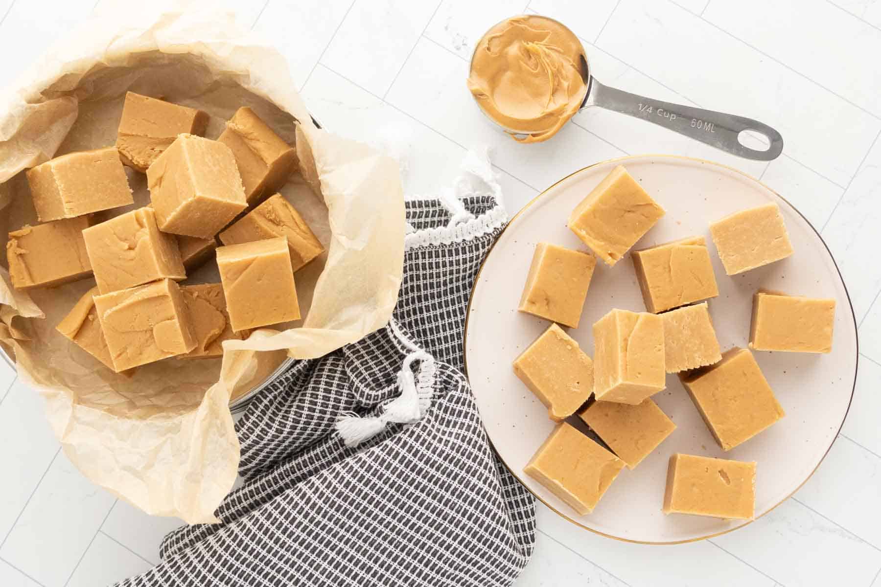 Squares of peanut butter fudge are arranged on a plate and in a parchment-lined bowl, with a measuring cup of peanut butter nearby on a white surface.