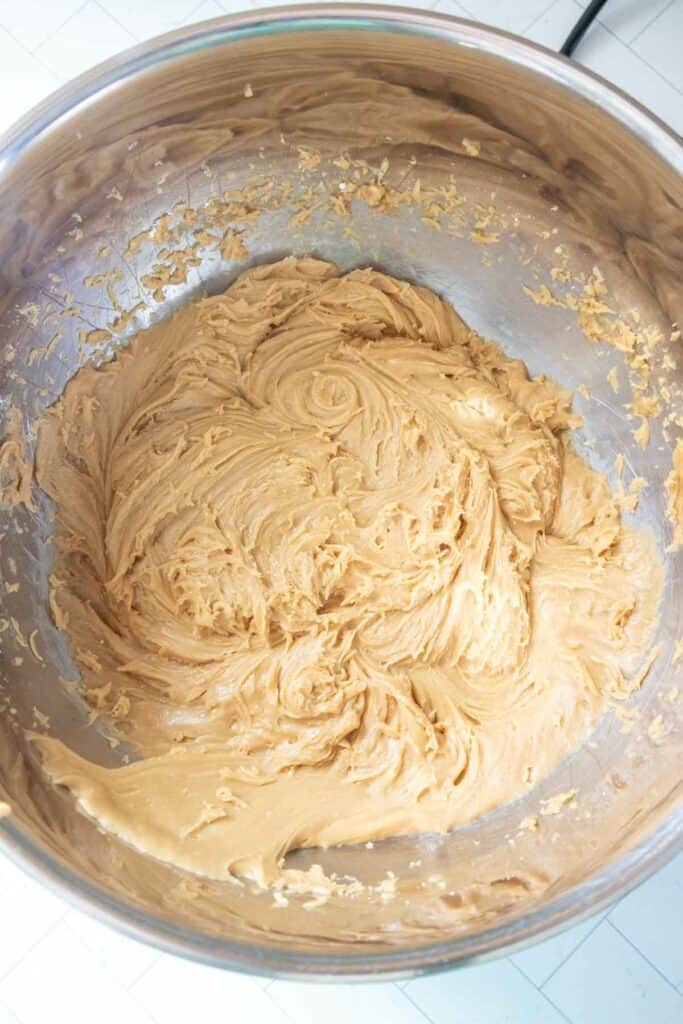A large metal mixing bowl filled with creamy, light brown batter, likely cookie dough or cake batter, on a white surface.