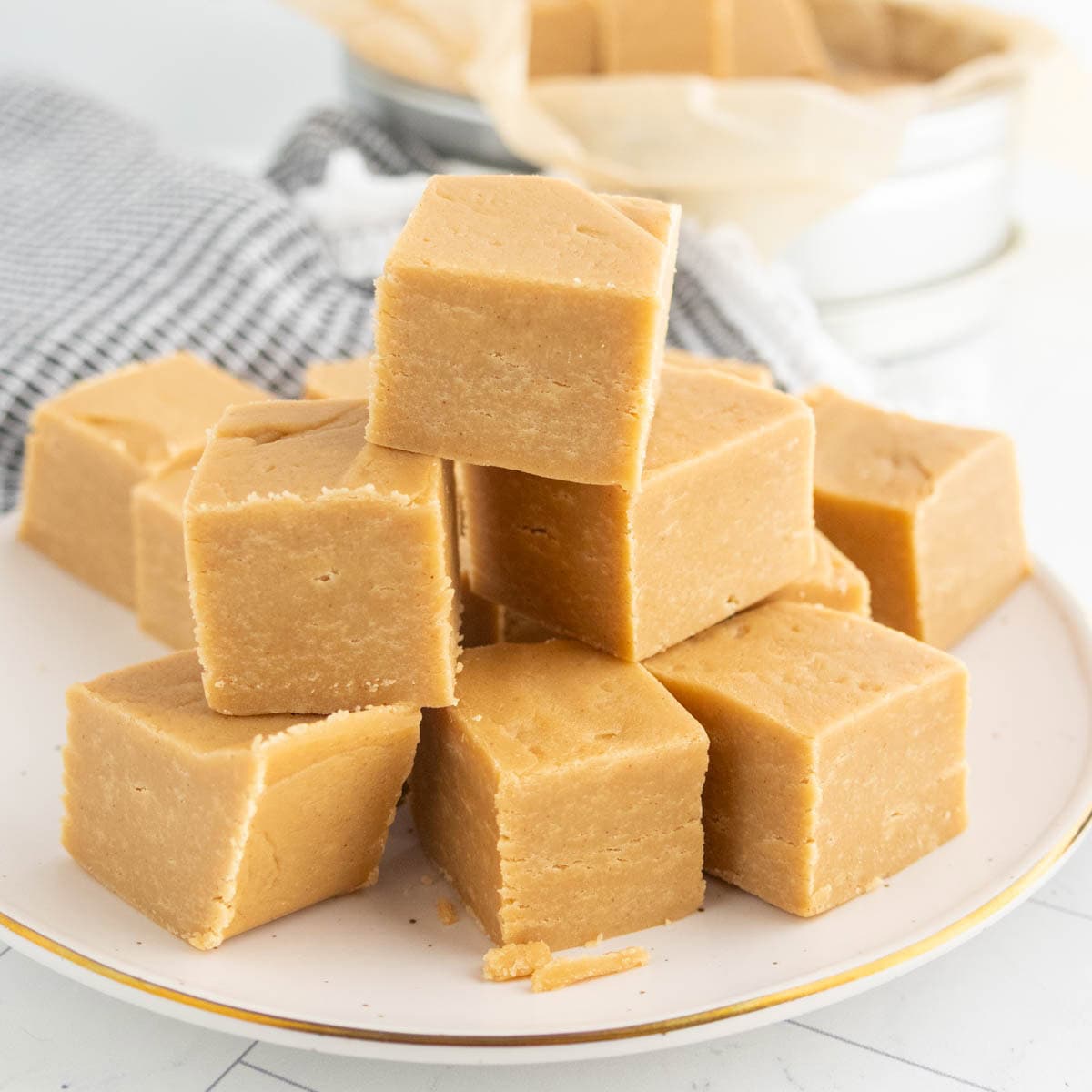 A plate stacked with square pieces of light brown fudge, arranged in a pyramid, with a cloth and additional fudge in the background.