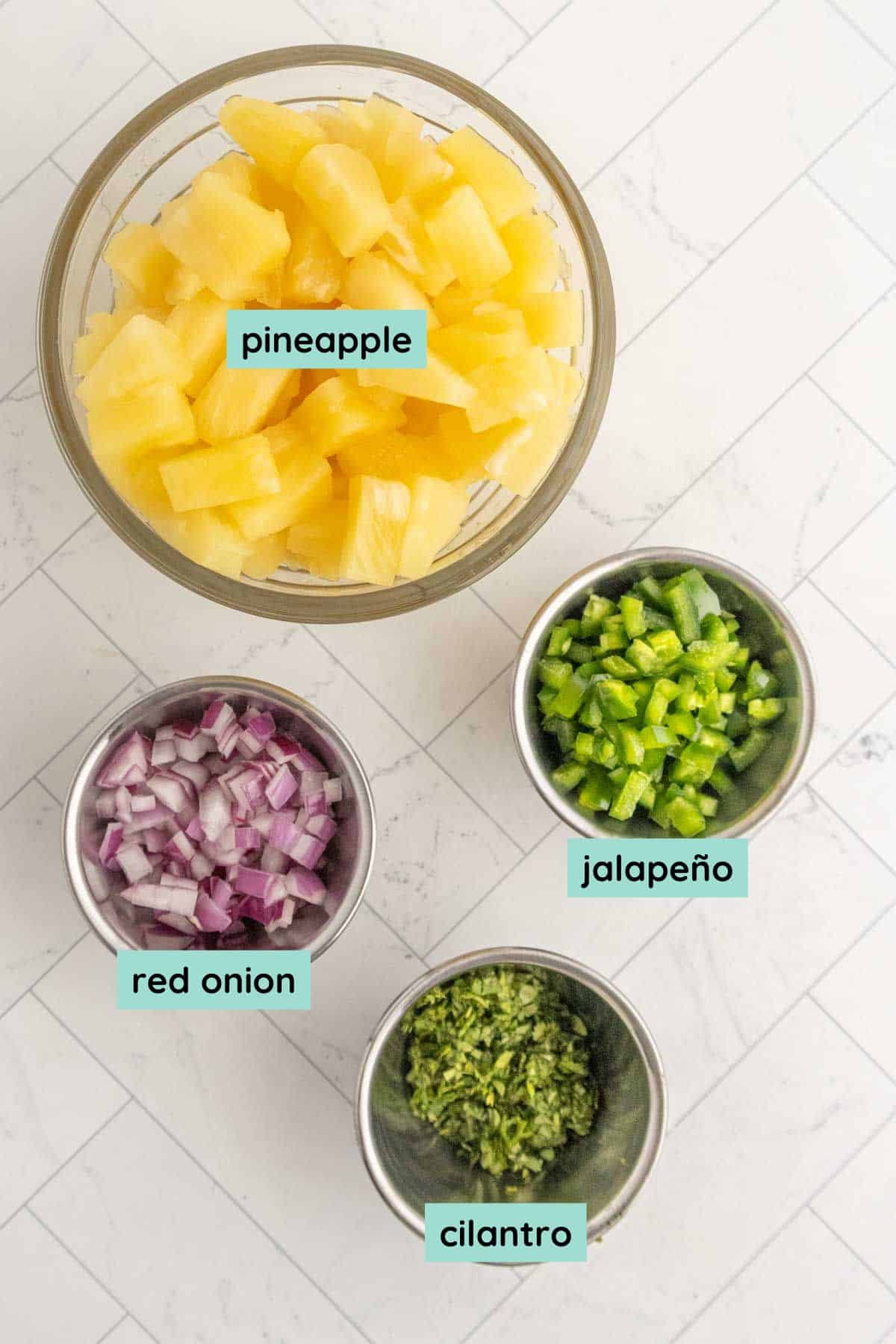 Four bowls on a white surface containing diced pineapple, chopped jalapeño, chopped red onion, and chopped cilantro, each labeled with text.