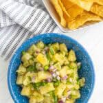 A blue bowl filled with pineapple salsa, featuring chopped pineapple, red onion, cilantro, and jalapeño, next to a basket of tortilla chips on a white surface.