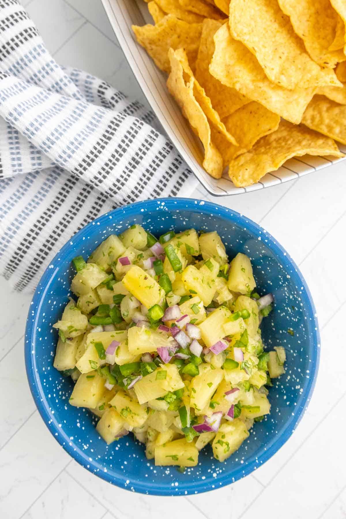 A blue bowl filled with pineapple salsa, featuring chopped pineapple, red onion, cilantro, and jalapeño, next to a basket of tortilla chips on a white surface.