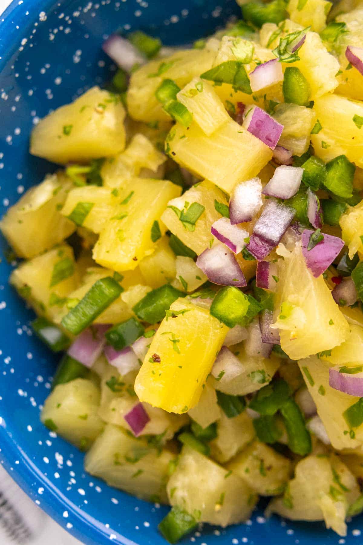 A close-up of pineapple salsa with diced pineapple, red onion, jalapeno, and cilantro in a blue speckled bowl.