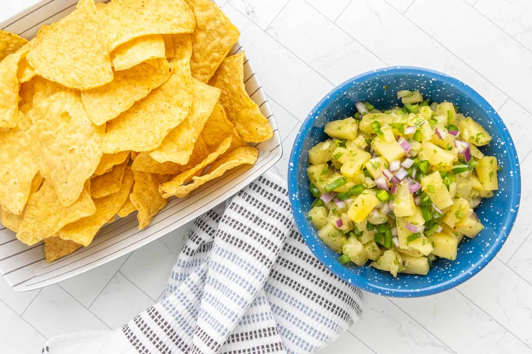 A basket of tortilla chips next to a blue bowl of pineapple salsa with chopped onions and herbs, placed on a white surface with a striped towel.