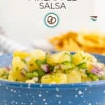 A blue bowl filled with pineapple salsa made of pineapple, red onion, and green herbs, with tortilla chips blurred in the background. Text on image reads "Pineapple Salsa".