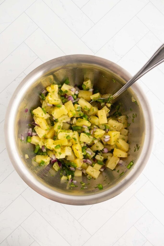 A metal bowl filled with a mixture of chopped pineapple, red onion, green pepper, and herbs, with a spoon inside.