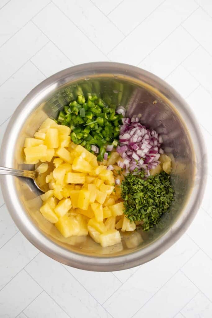 A metal bowl containing chopped pineapple, diced green bell pepper, chopped red onion, and chopped cilantro, with a spoon inside.