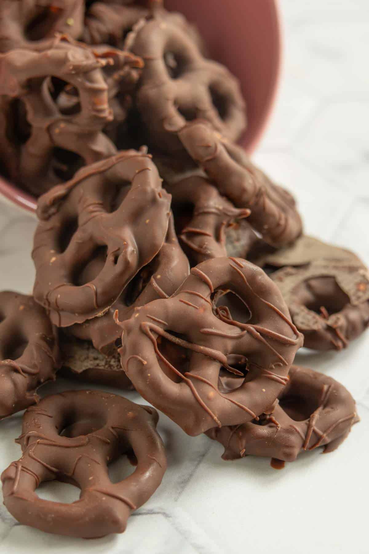 Chocolate-covered pretzels spilling out of a pink bowl onto a white surface.