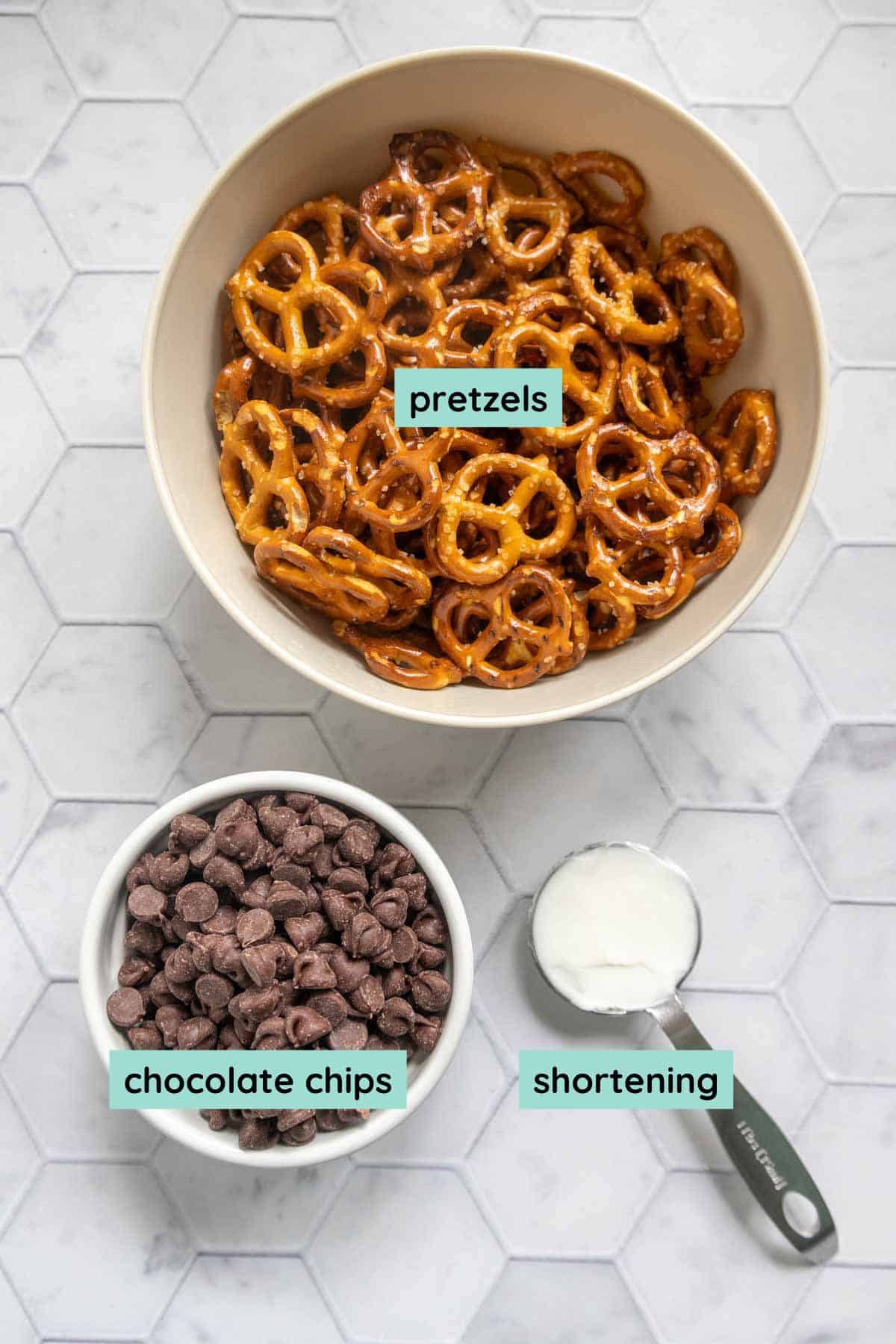 A bowl of pretzels, a bowl of chocolate chips, and a measuring cup of shortening on a white hexagon-tile surface; each item is labeled.