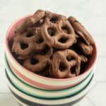 A bowl filled with chocolate-covered pretzels, placed on top of a stack of striped ceramic bowls on a light-colored surface.