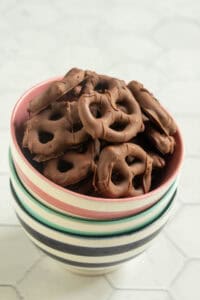 A bowl filled with chocolate-covered pretzels, placed on top of a stack of striped ceramic bowls on a light-colored surface.