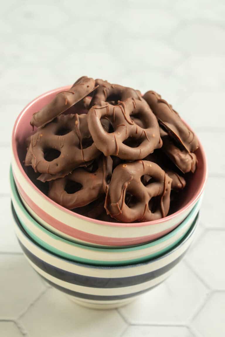A bowl filled with chocolate-covered pretzels, placed on top of a stack of striped ceramic bowls on a light-colored surface.