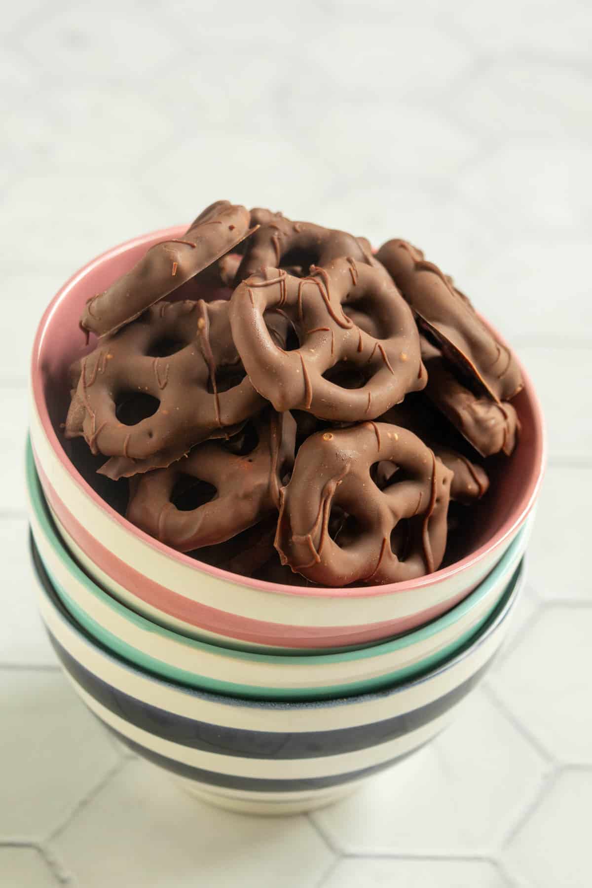 A bowl filled with chocolate-covered pretzels, placed on top of a stack of striped ceramic bowls on a light-colored surface.