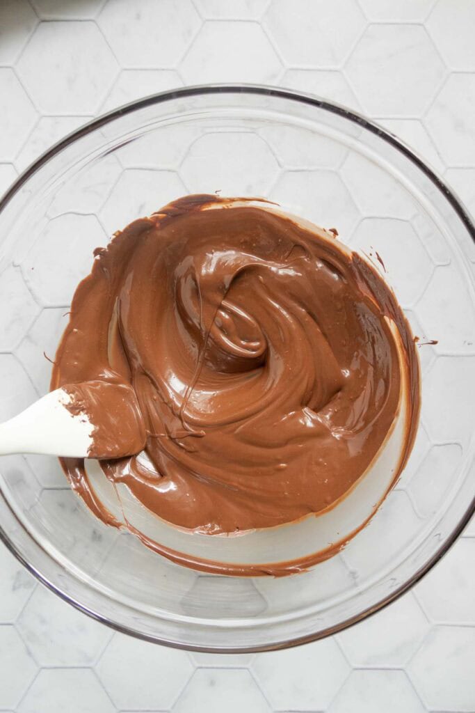 A clear glass bowl filled with melted chocolate being stirred by a white spatula, on a white hexagonal tile surface.