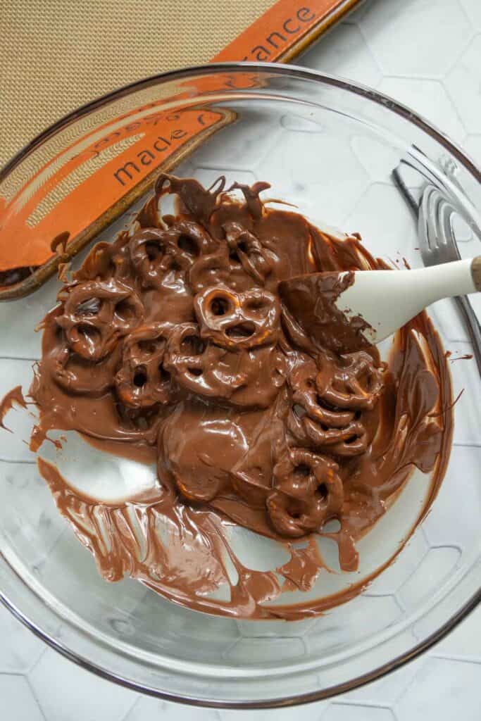 A glass bowl filled with pretzels coated in melted chocolate being mixed with a white spatula.