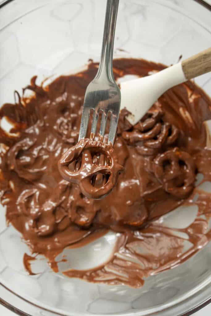 A fork holding a pretzel coated in melted chocolate above a bowl filled with more chocolate-covered pretzels and a white spatula.
