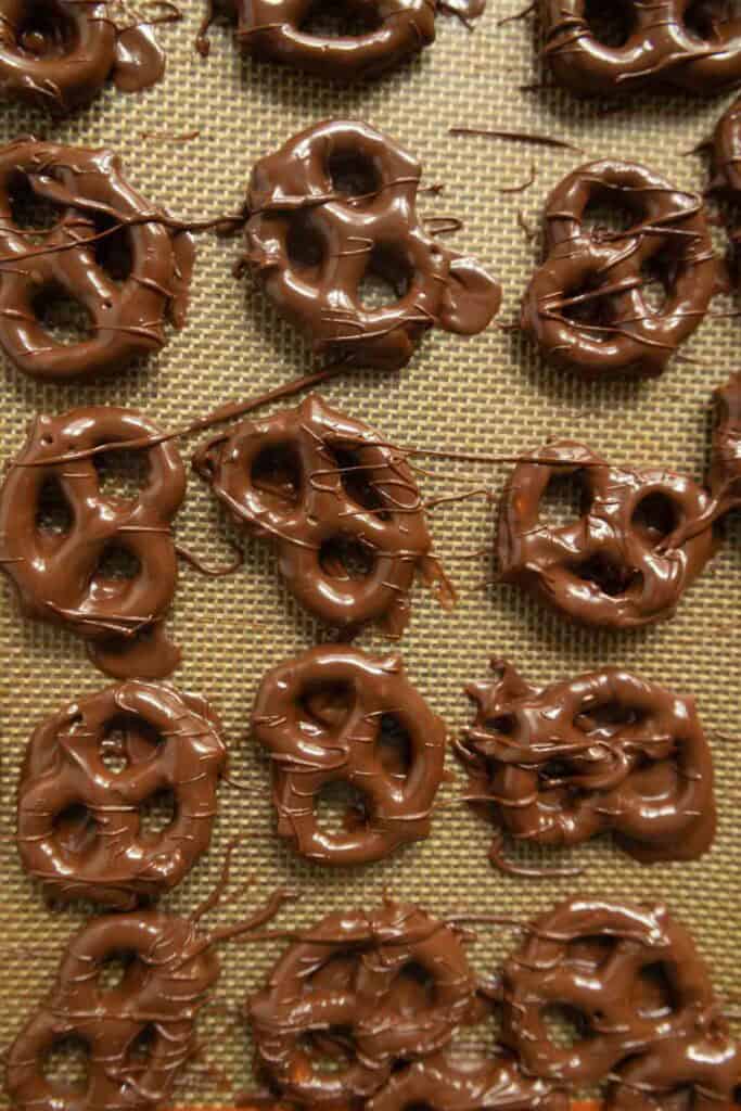 Chocolate-covered pretzels cooling on a non-stick baking mat, arranged in rows with some chocolate drizzled and pooled around the edges.