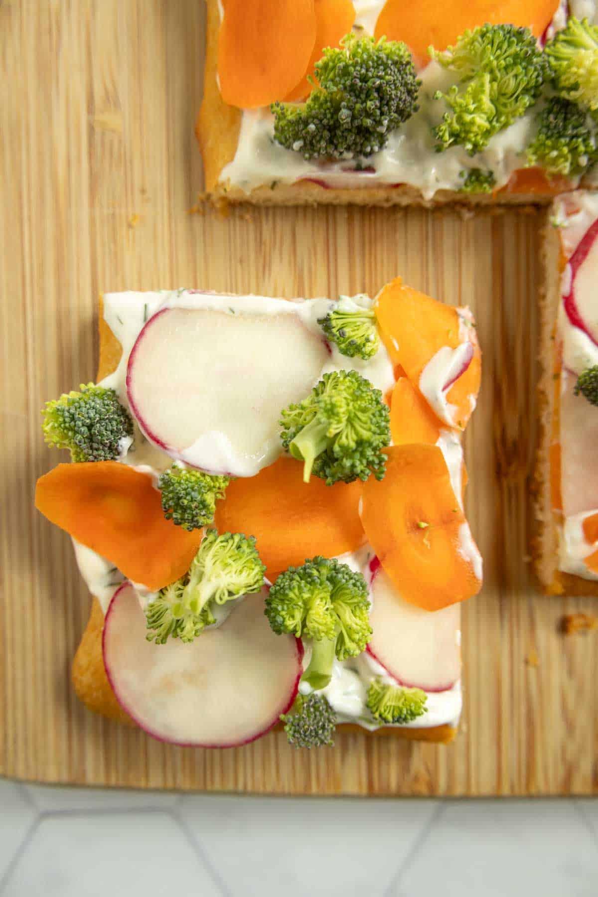 A square piece of vegetable-topped flatbread with cream cheese, sliced radishes, carrots, and broccoli on a wooden board.