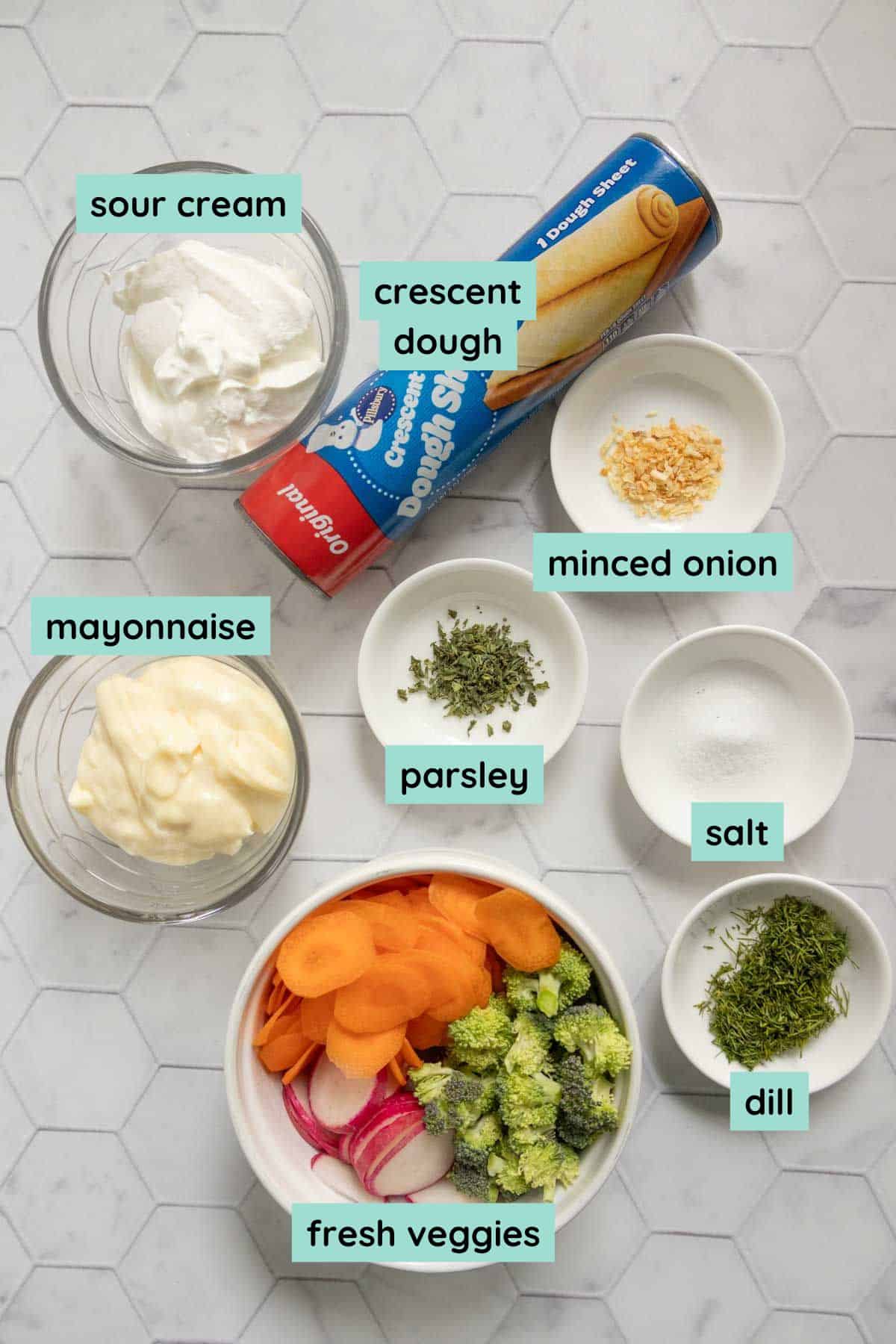 Ingredients for a recipe arranged on a countertop, including sour cream, crescent dough, mayonnaise, minced onion, parsley, salt, dill, and fresh sliced carrots and broccoli.