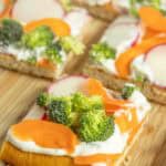 Square pieces of flatbread topped with cream cheese, sliced carrots, broccoli florets, and radishes on a wooden surface.