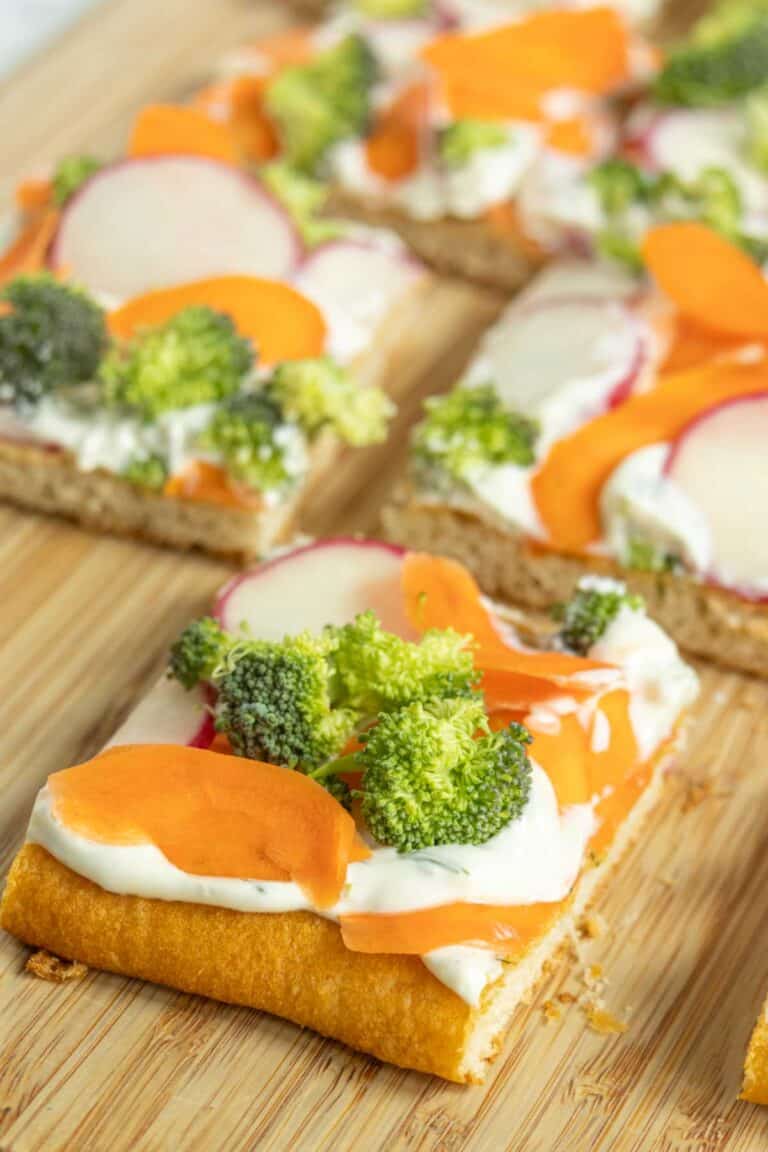 Square pieces of flatbread topped with cream cheese, sliced carrots, broccoli florets, and radishes on a wooden surface.