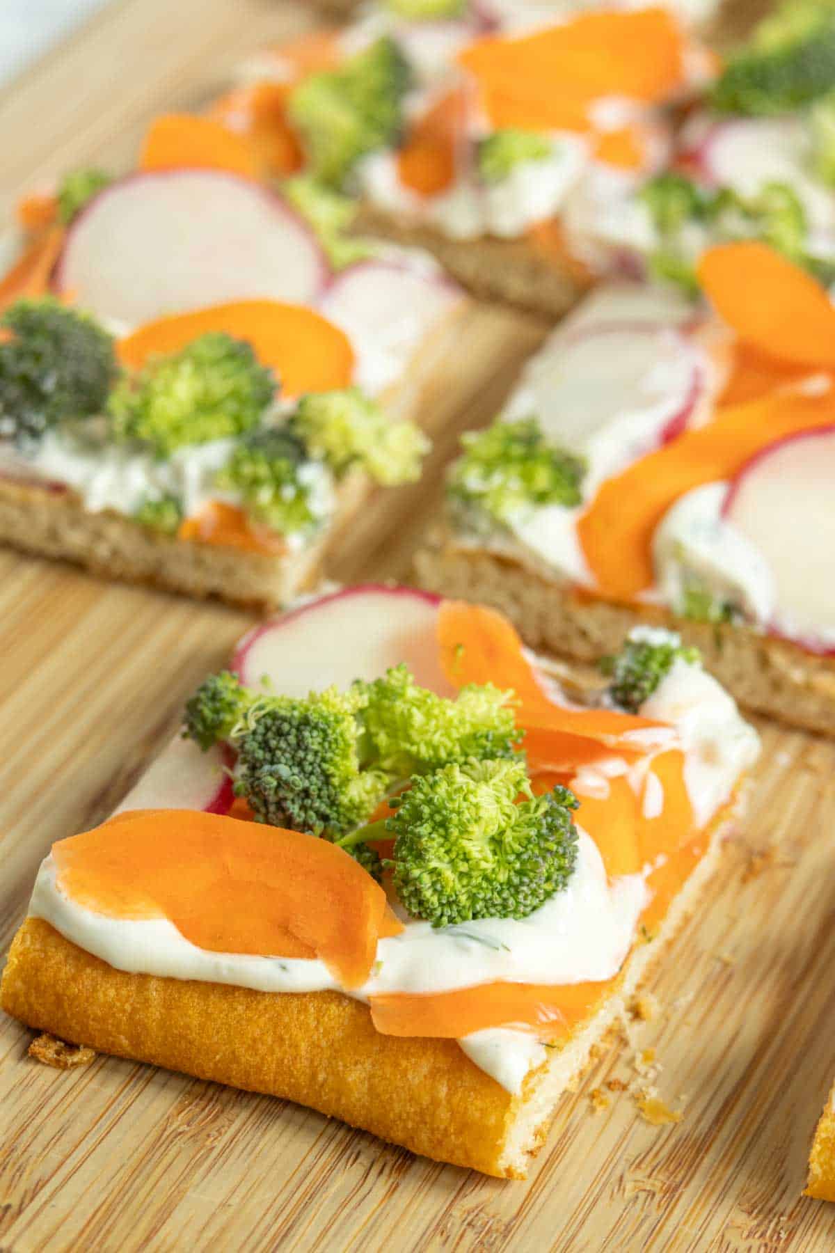 Square pieces of flatbread topped with cream cheese, sliced carrots, broccoli florets, and radishes on a wooden surface.