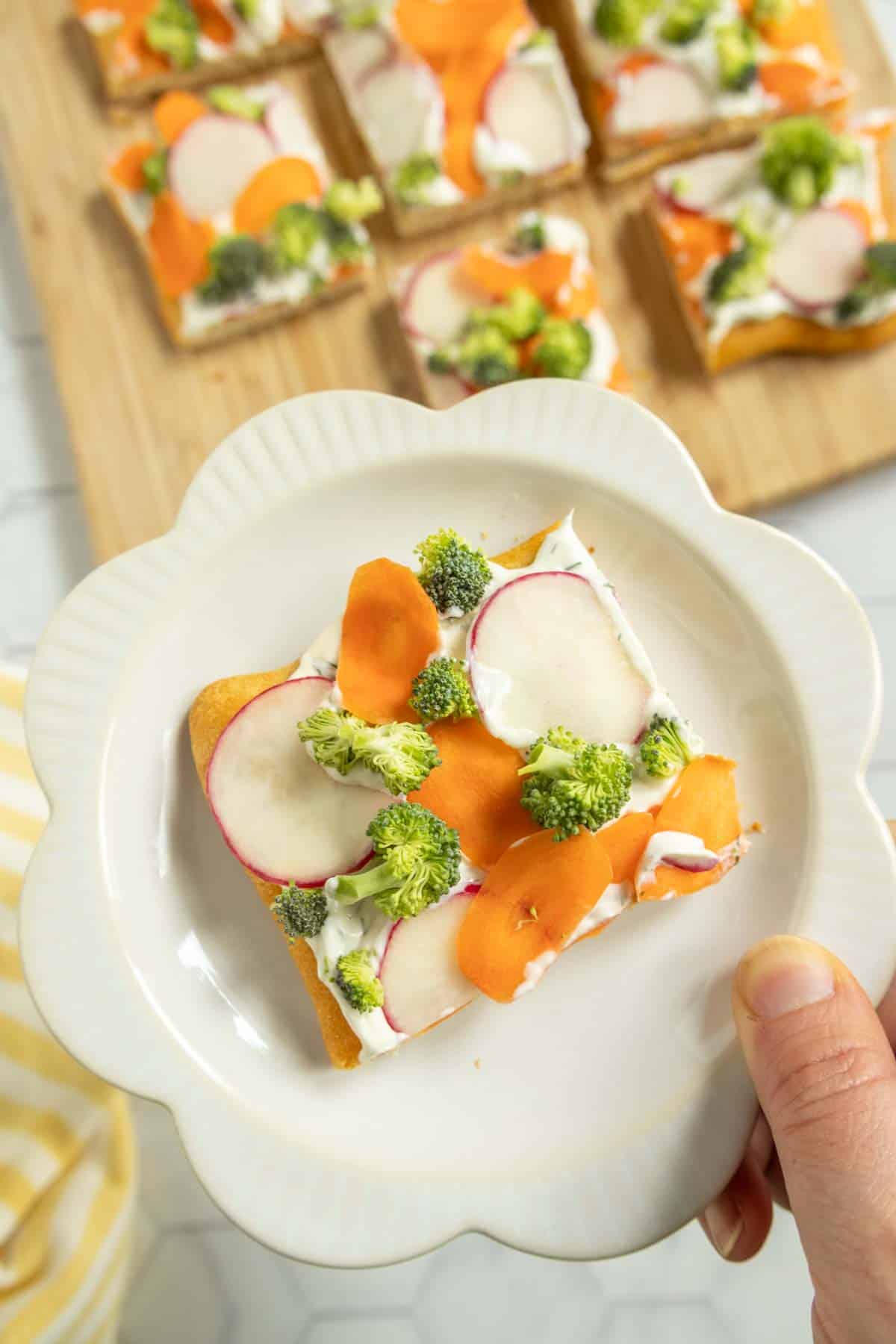 A hand holds a plate with a square slice of veggie pizza topped with cream cheese, carrot, radish, and broccoli; more slices are on a wooden board in the background.