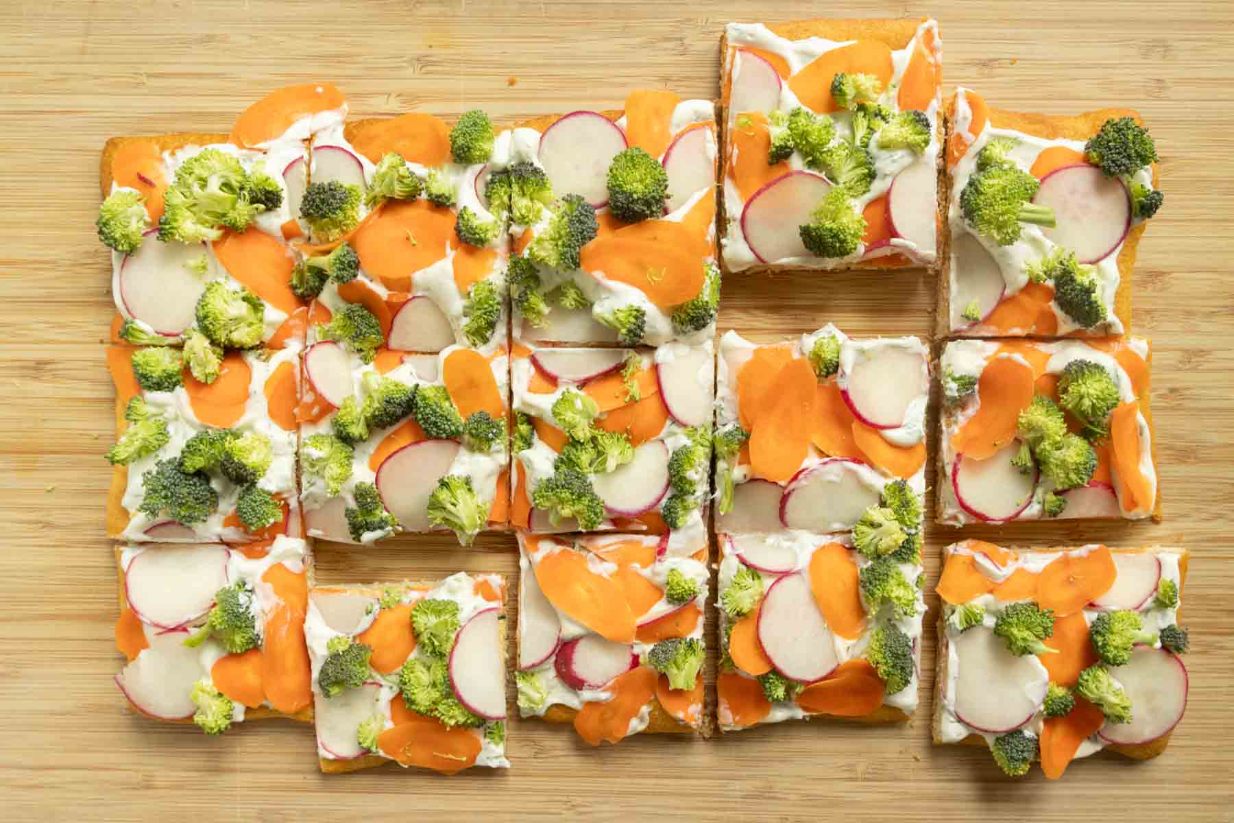 Rectangular veggie pizza cut into squares, topped with broccoli, radish, and carrot slices on a wooden surface.