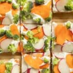 Rectangular slices of veggie pizza appetizer topped with broccoli, radish, and carrot on a wooden surface. The website happysnackcidents.com is displayed at the bottom.