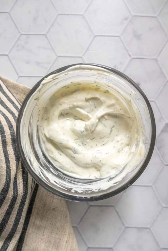 A glass bowl filled with creamy white ranch dressing, speckled with herbs, sits on a hexagon-tiled surface next to a striped cloth napkin.
