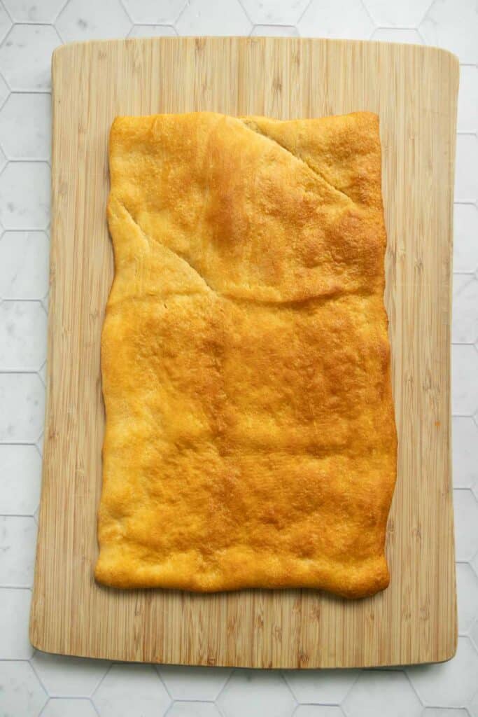 Rectangular sheet of baked golden-brown dough on a wooden cutting board, placed on a white hexagonal tile surface.
