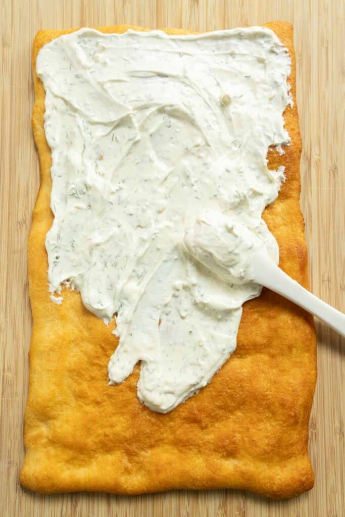 A baked rectangular crust on a wooden surface being spread with a layer of herbed cream cheese using a white spatula.