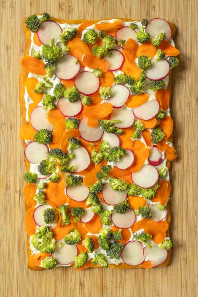 A rectangular flatbread topped with cream cheese, sliced radishes, carrots, and broccoli on a wooden surface.