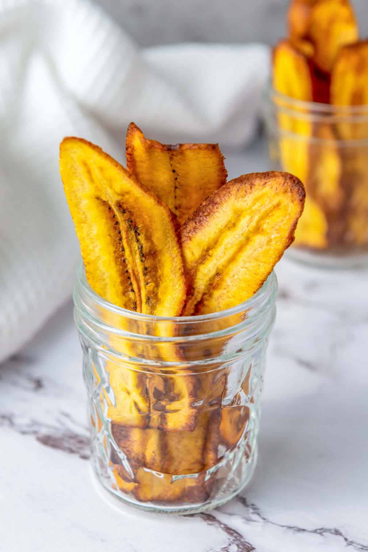 Glass jar filled with several crispy, golden-brown plantain chips on a white marble surface, with another jar blurred in the background.