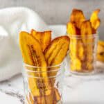 Two glass jars filled with baked plantain chips are placed on a marble surface with a white cloth and another jar in the background.