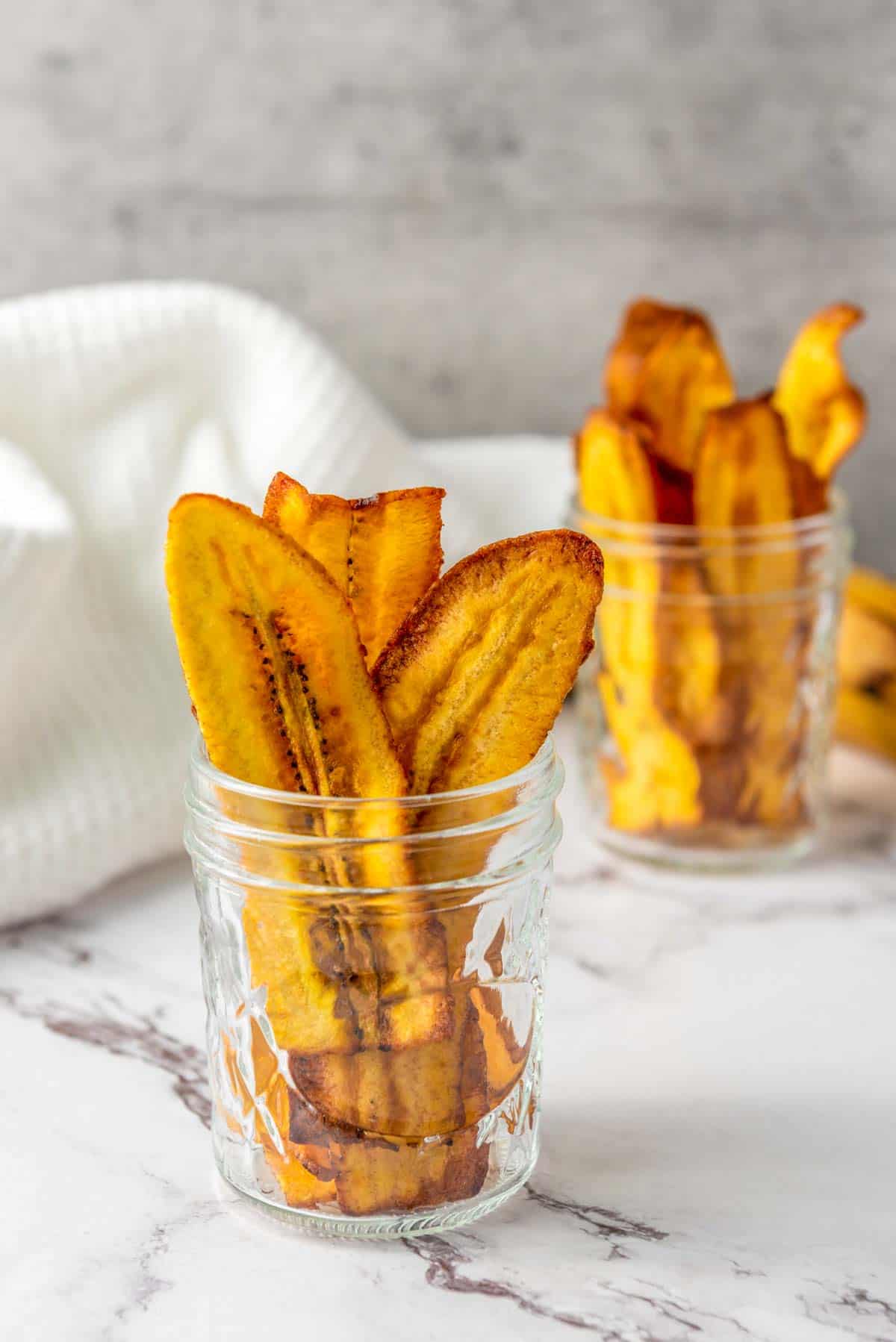 Two glass jars filled with baked plantain chips are placed on a marble surface with a white cloth and another jar in the background.