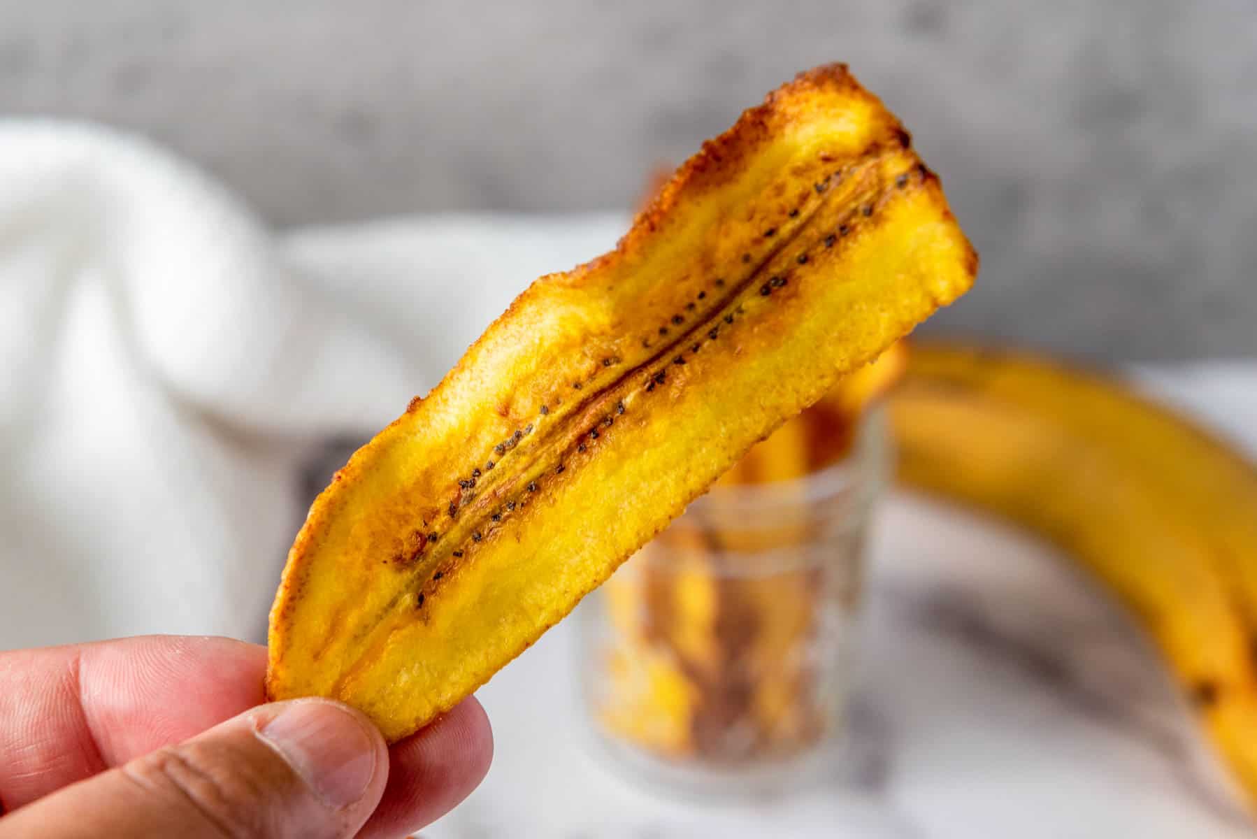 A hand holds a single yellow plantain chip. In the background, a glass jar with more chips and a whole plantain are visible on a light surface.