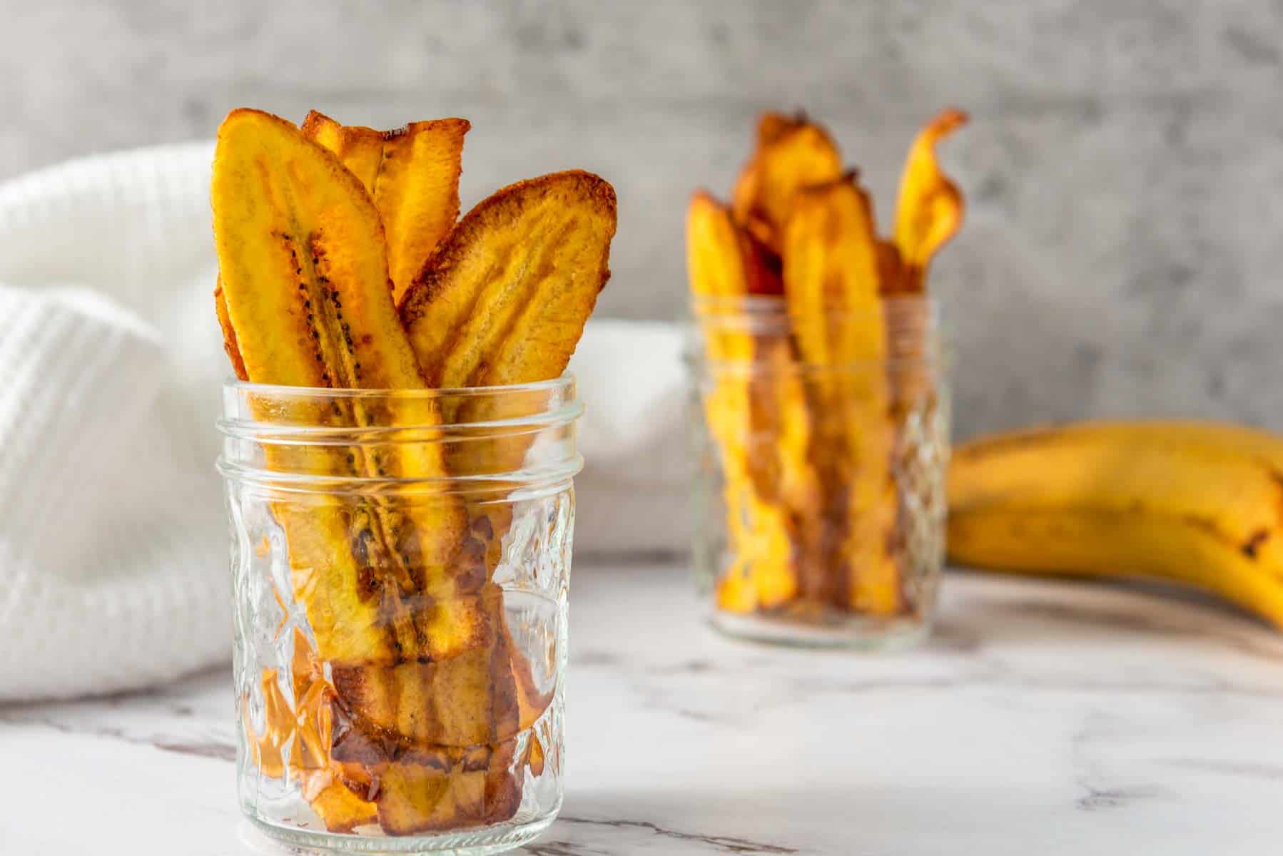 Two glass jars filled with baked plantain chips sit on a marble surface, with a banana and a white cloth in the background.