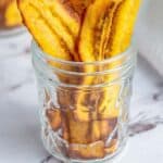 Glass jar filled with golden-brown fried plantain chips, placed on a marble surface. Text at the bottom reads "Fried Plantain Chips happysnackcidents.com".