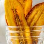 Several crispy plantain chips stand upright in a clear glass jar, with text at the bottom reading "PLANTAIN CHIPS" and a website URL.