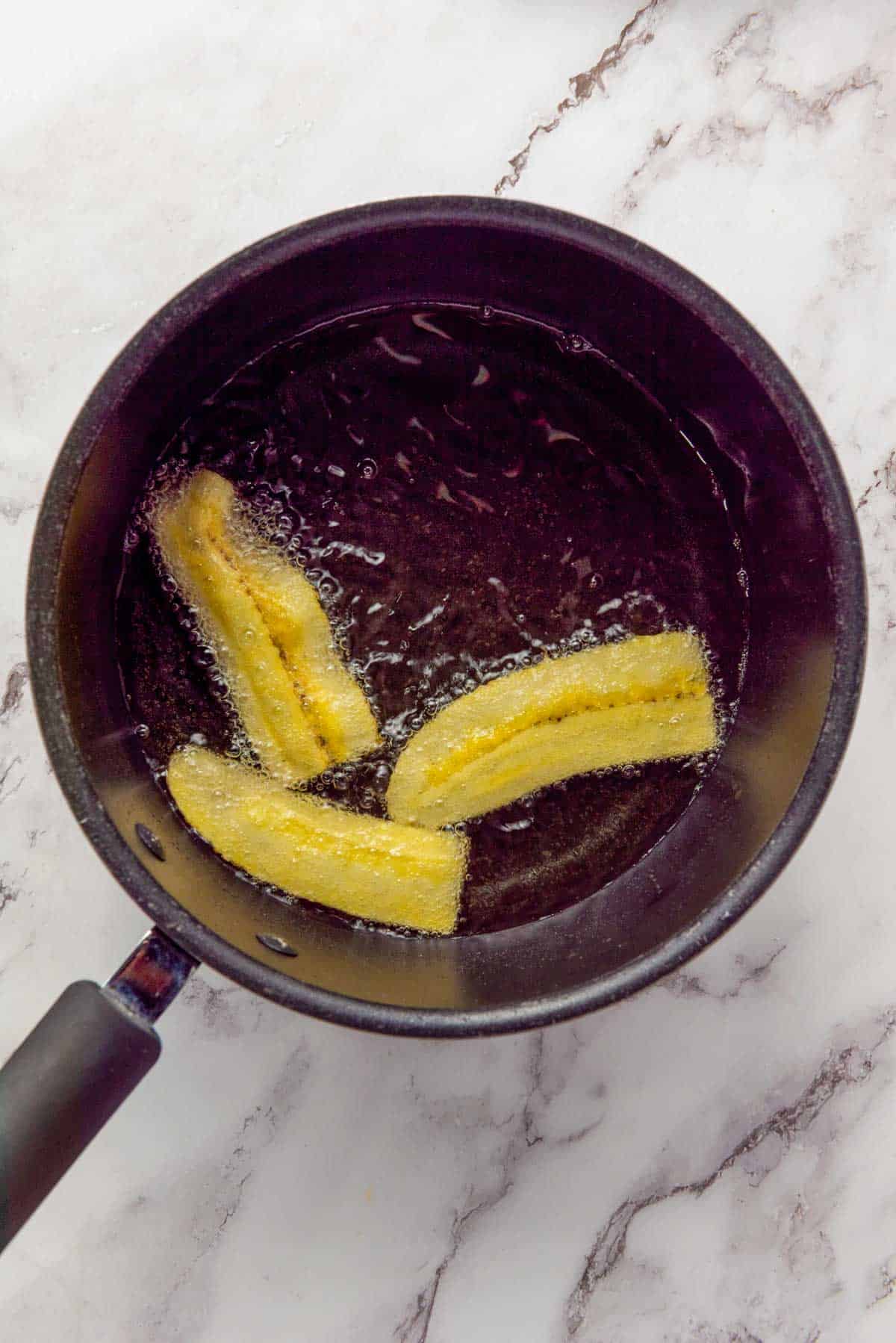 Three plantain slices frying in hot oil inside a black pan on a marble countertop.