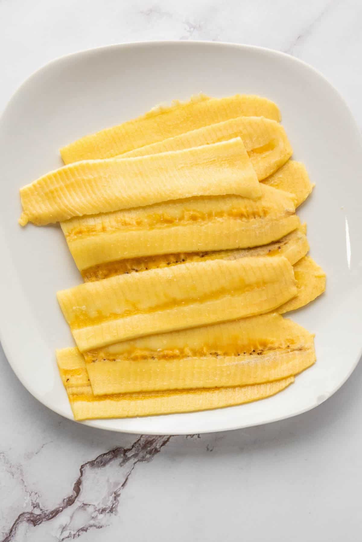 Several thin slices of raw plantain are arranged on a white plate, which is set on a white marble surface.