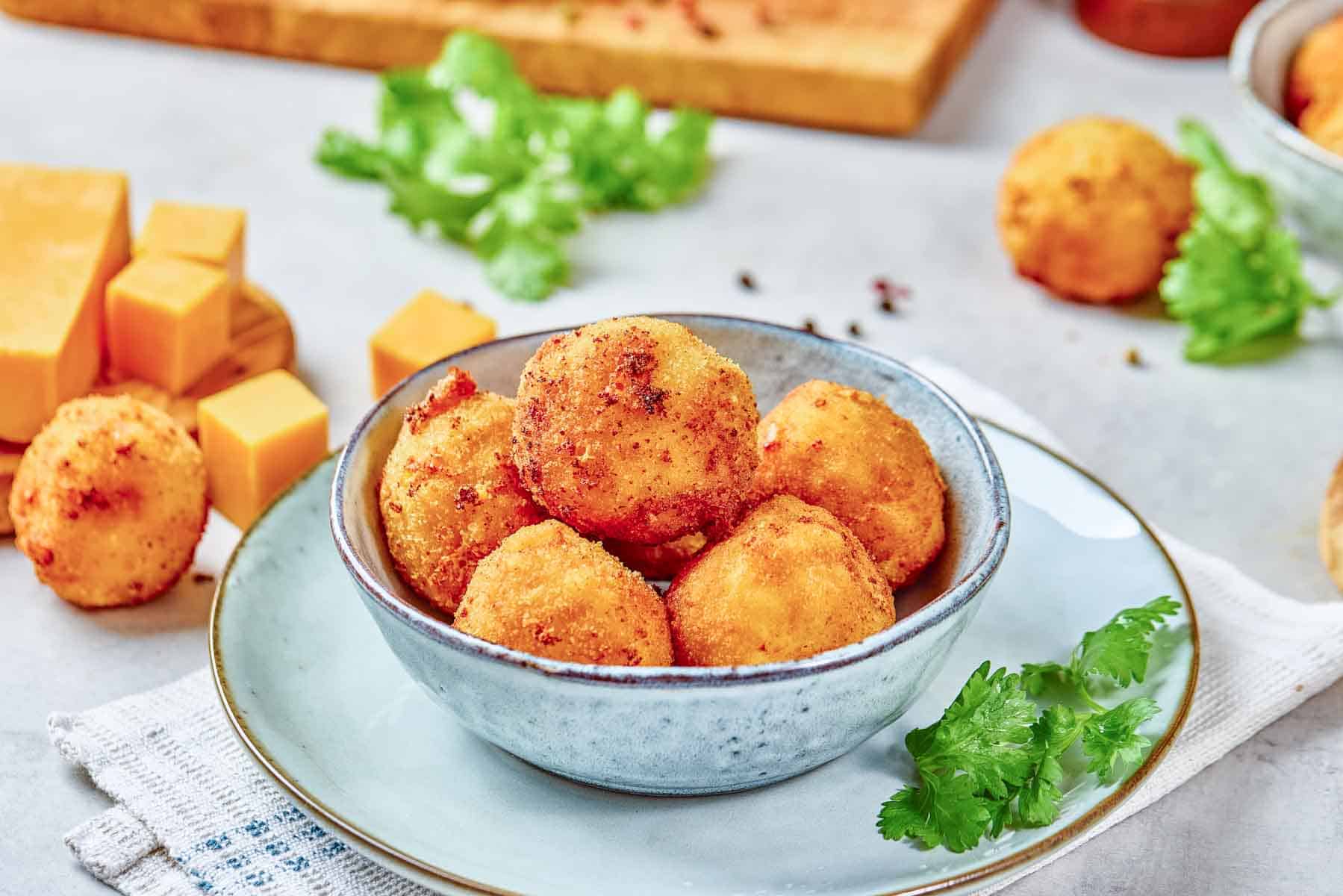 A bowl of golden, breaded cheese balls sits on a plate, surrounded by fresh herbs and cubes of cheddar cheese on a light background.