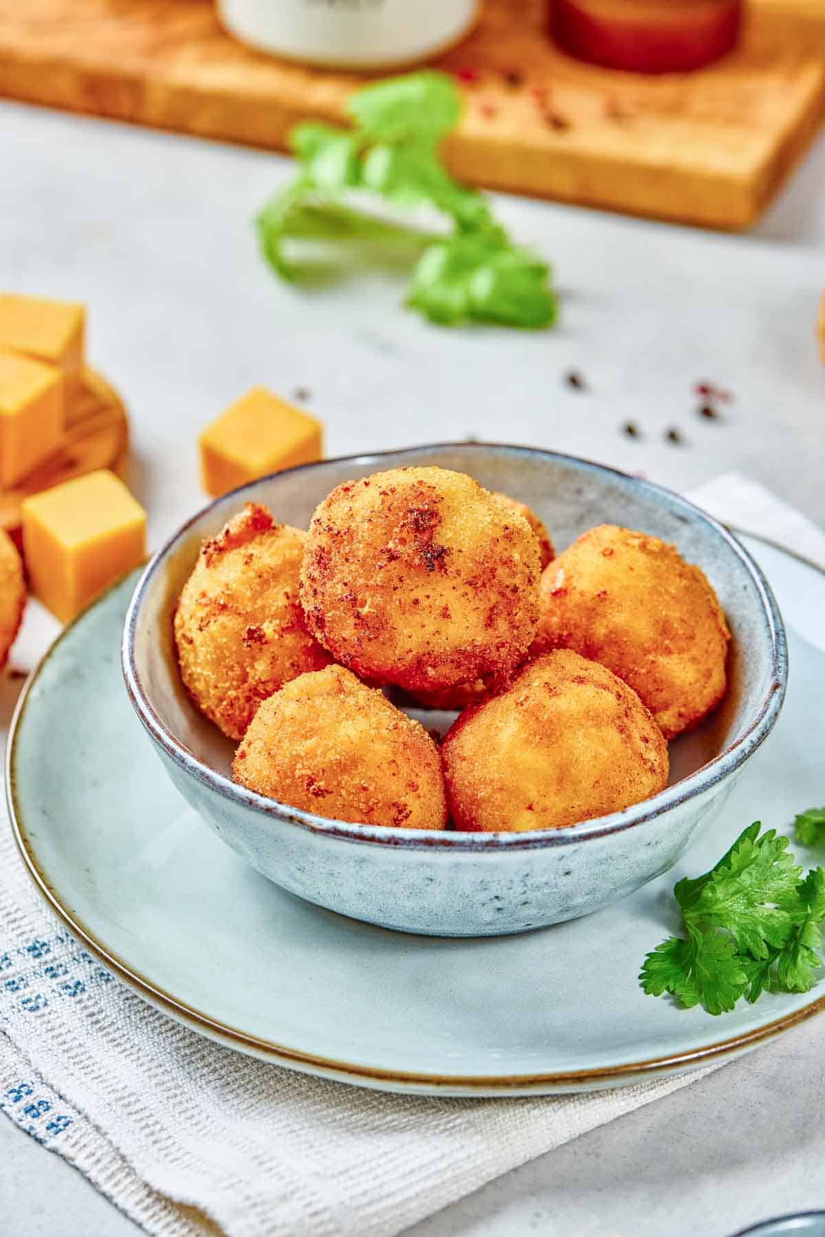 A bowl with five golden-brown breaded cheese balls on a plate, with cubed cheese and fresh cilantro nearby on the table.