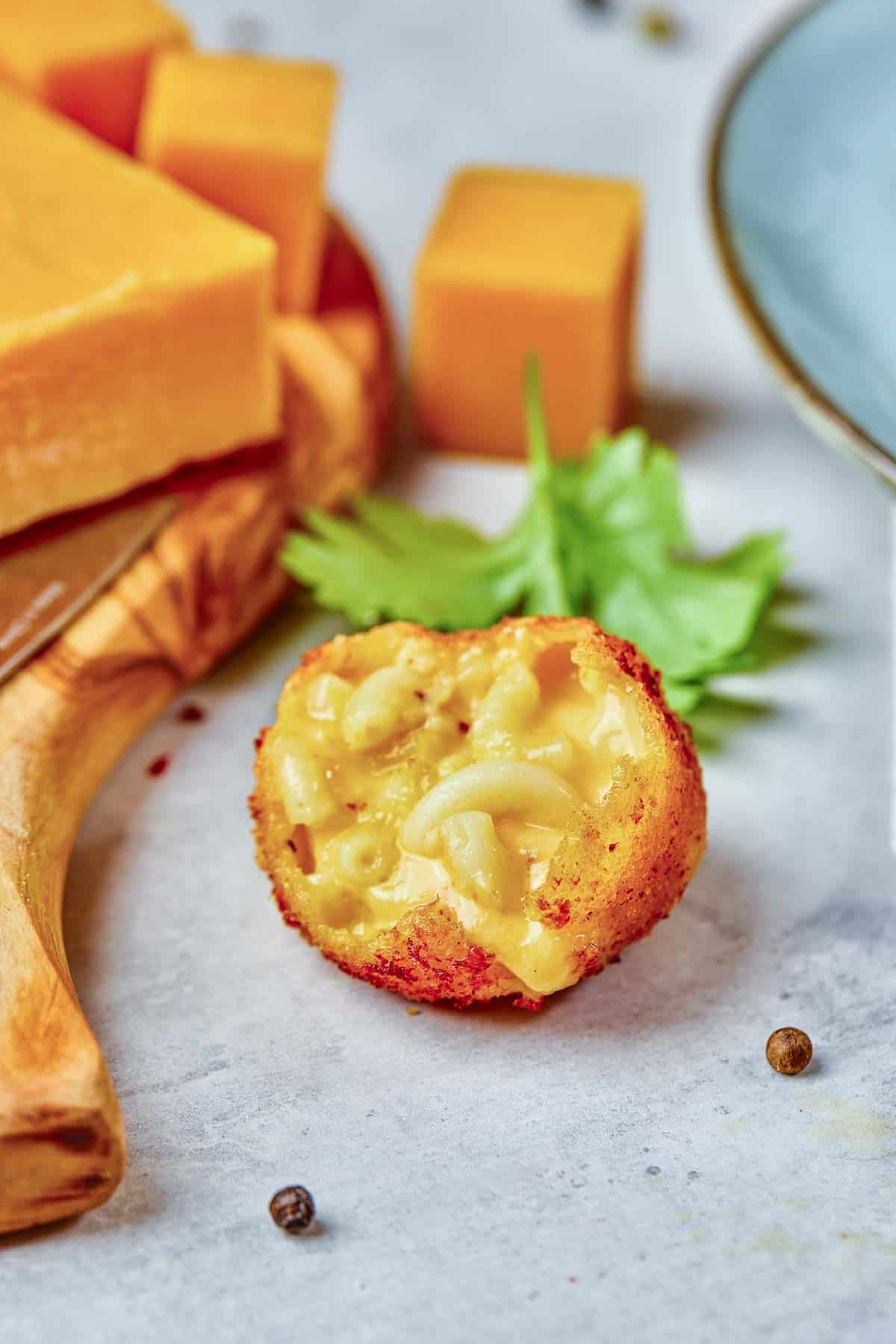 A breaded, fried macaroni and cheese ball is cut open, showing the creamy pasta inside. Cheddar cheese cubes, parsley, and a plate are nearby on a light surface.